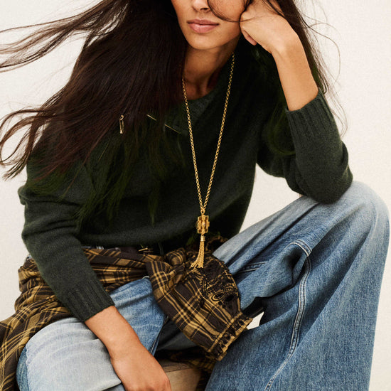 A woman posing in a dark green knit sweater, wide-leg blue jeans, a brown plaid shirt tied around her waist, and a long gold pendant necklace against a plain off-white background.