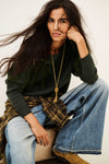 A woman posing in a dark green knit sweater, wide-leg blue jeans, a brown plaid shirt tied around her waist, and a long gold pendant necklace against a plain off-white background.