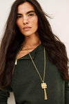 A woman with long dark hair wearing a dark green zip-up sweater and a gold chain necklace with a square pendant and tassel, photographed against a plain light background.