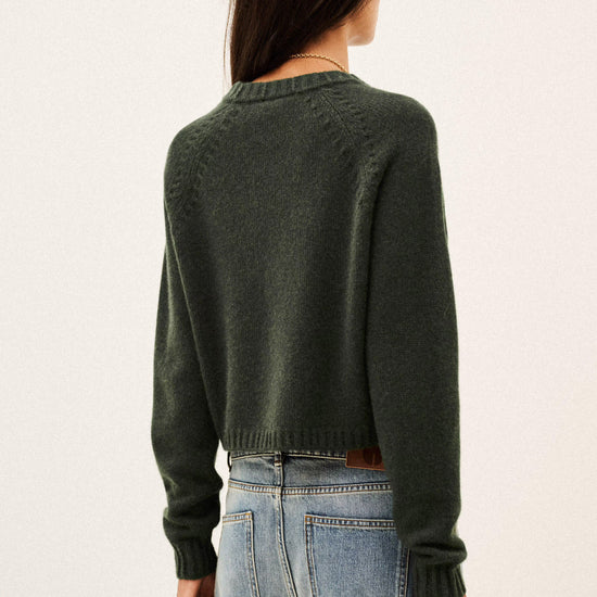 A woman viewed from the back wearing a dark olive green knit sweater and light blue jeans against a plain off-white background.
