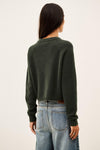 A woman viewed from the back wearing a dark olive green knit sweater and light blue jeans against a plain off-white background.