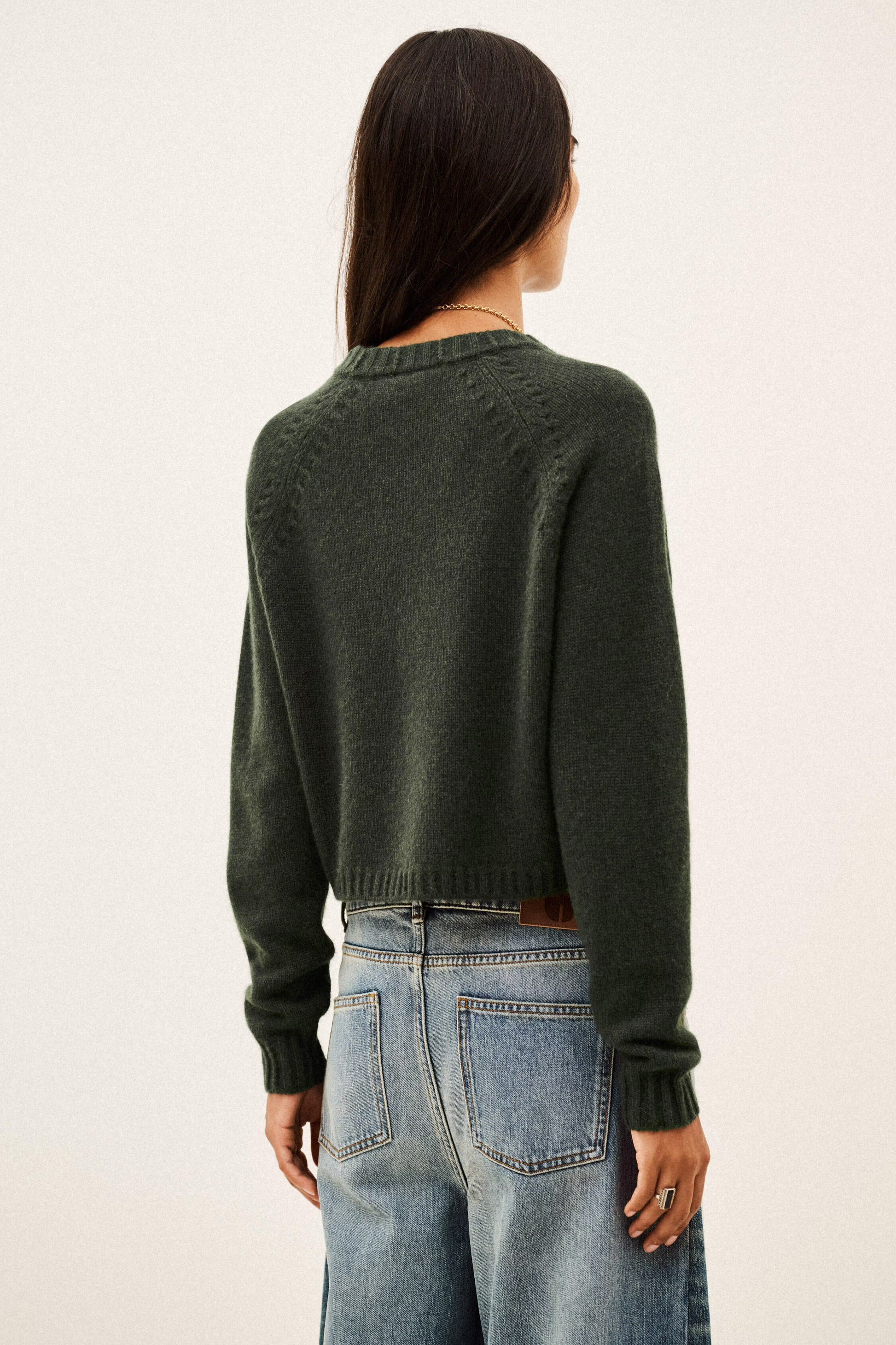 A woman viewed from the back wearing a dark olive green knit sweater and light blue jeans against a plain off-white background.