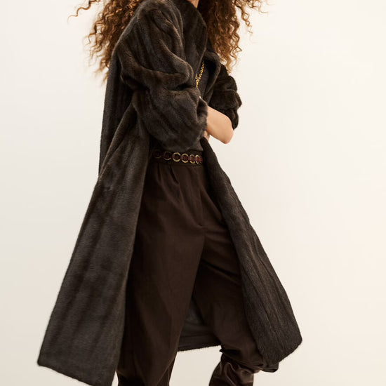 A model with voluminous curly brown hair is wearing a dark brown textured long coat, matching dark brown trousers, a patterned belt, and dark brown heeled boots against a plain off-white studio background.