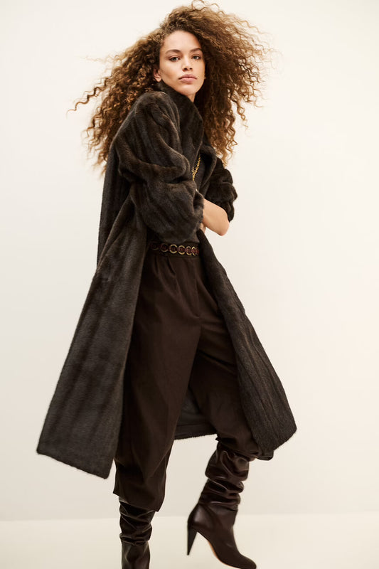 A model with voluminous curly brown hair is wearing a dark brown textured long coat, matching dark brown trousers, a patterned belt, and dark brown heeled boots against a plain off-white studio background.