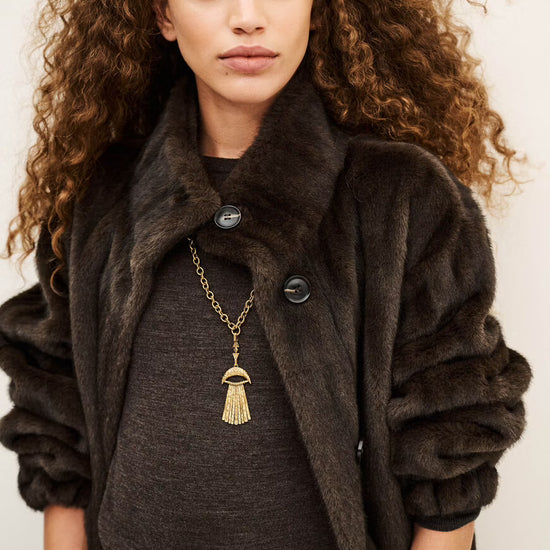 A model with voluminous curly brown hair wearing a dark brown faux fur coat over a charcoal gray top, accessorized with a gold tassel pendant necklace and a brown chain belt.