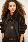 A model with voluminous curly brown hair wearing a dark brown faux fur coat over a charcoal gray top, accessorized with a gold tassel pendant necklace and a brown chain belt.