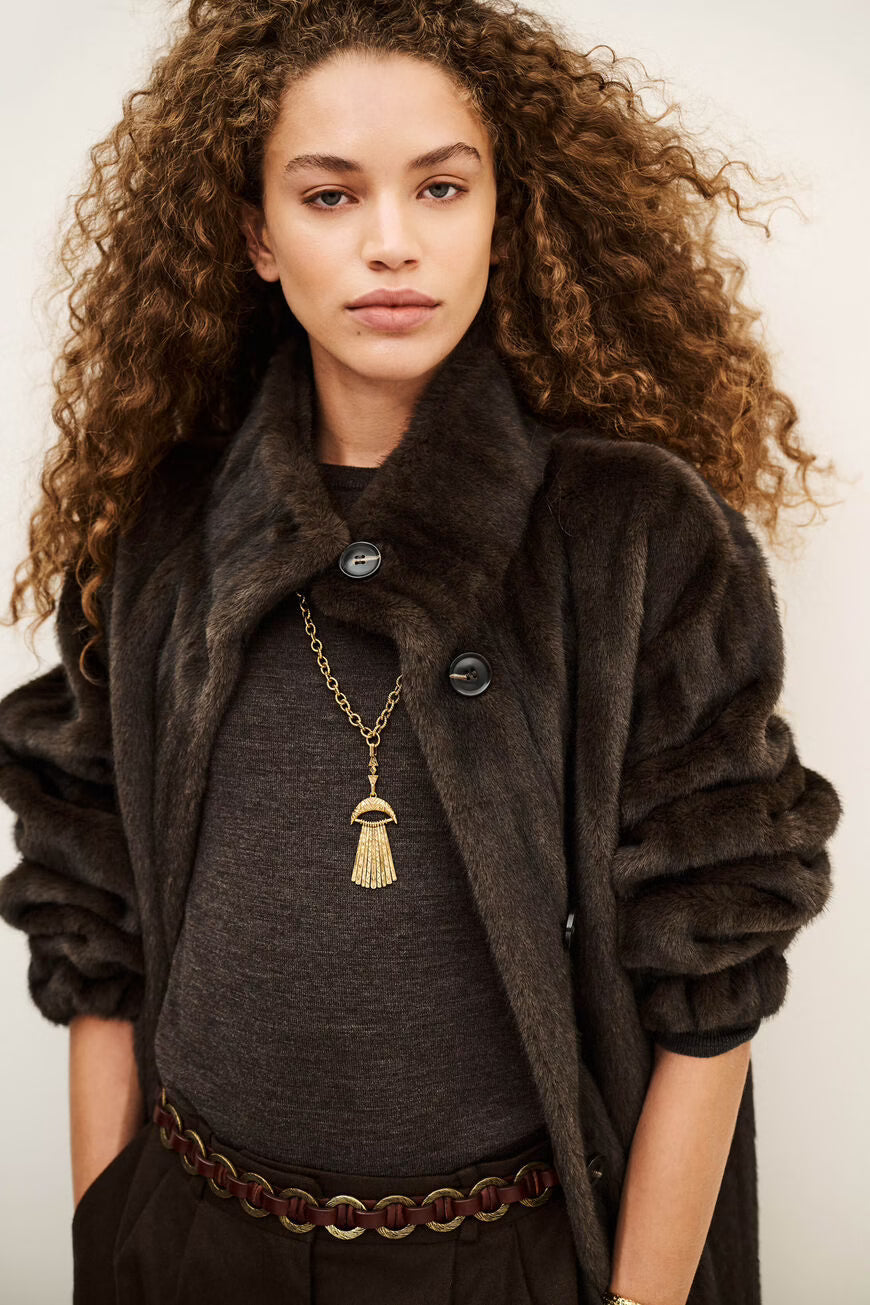 A model with voluminous curly brown hair wearing a dark brown faux fur coat over a charcoal gray top, accessorized with a gold tassel pendant necklace and a brown chain belt.