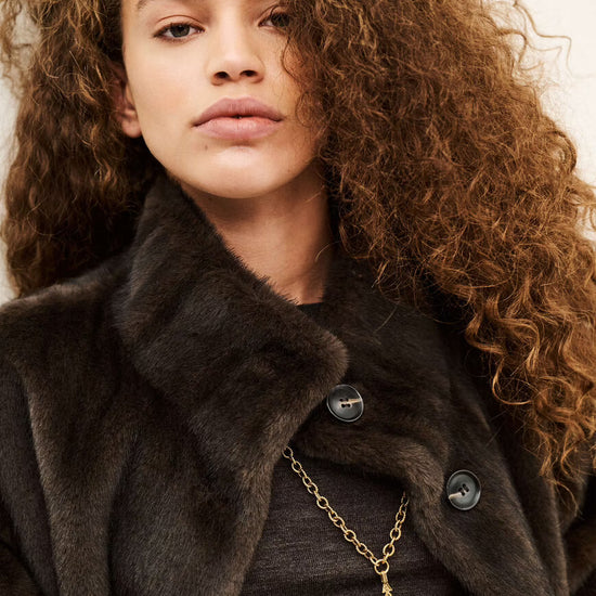 A close-up portrait of a model with voluminous curly brown hair wearing a dark brown fur-trimmed coat over a dark top, accessorized with a gold chain necklace featuring a tassel pendant.