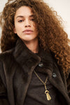 A close-up portrait of a model with voluminous curly brown hair wearing a dark brown fur-trimmed coat over a dark top, accessorized with a gold chain necklace featuring a tassel pendant.