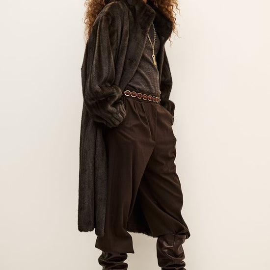 A model with curly brown hair is shown wearing a dark brown textured coat, matching dark brown trousers, a patterned belt, and dark brown heeled boots against a plain off-white studio background.