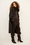 A model with curly brown hair is shown wearing a dark brown textured coat, matching dark brown trousers, a patterned belt, and dark brown heeled boots against a plain off-white studio background.