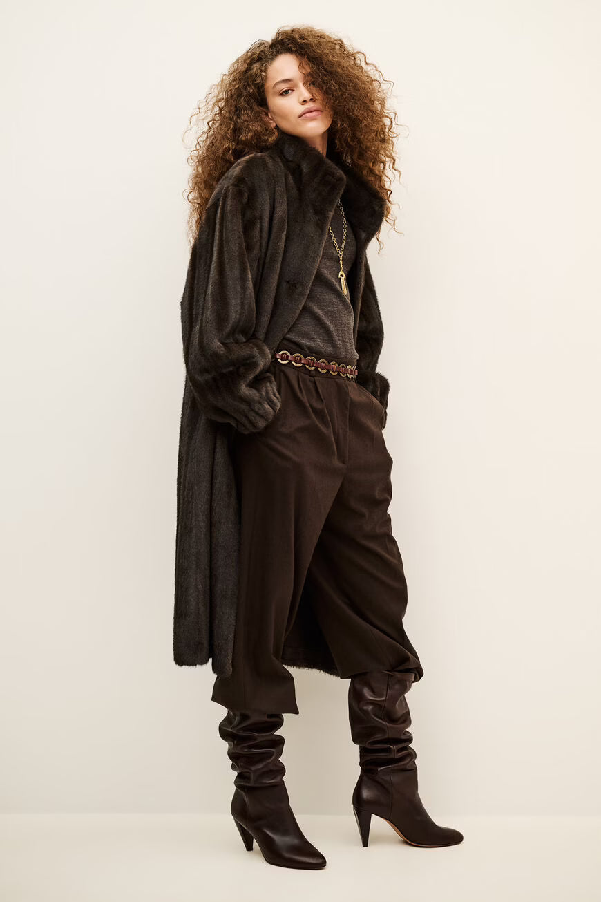 A model with curly brown hair is shown wearing a dark brown textured coat, matching dark brown trousers, a patterned belt, and dark brown heeled boots against a plain off-white studio background.
