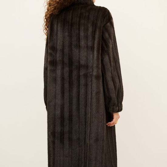 A model viewed from behind wearing a long, dark brown shaggy faux fur coat, black pants, and black boots against a plain off-white background.