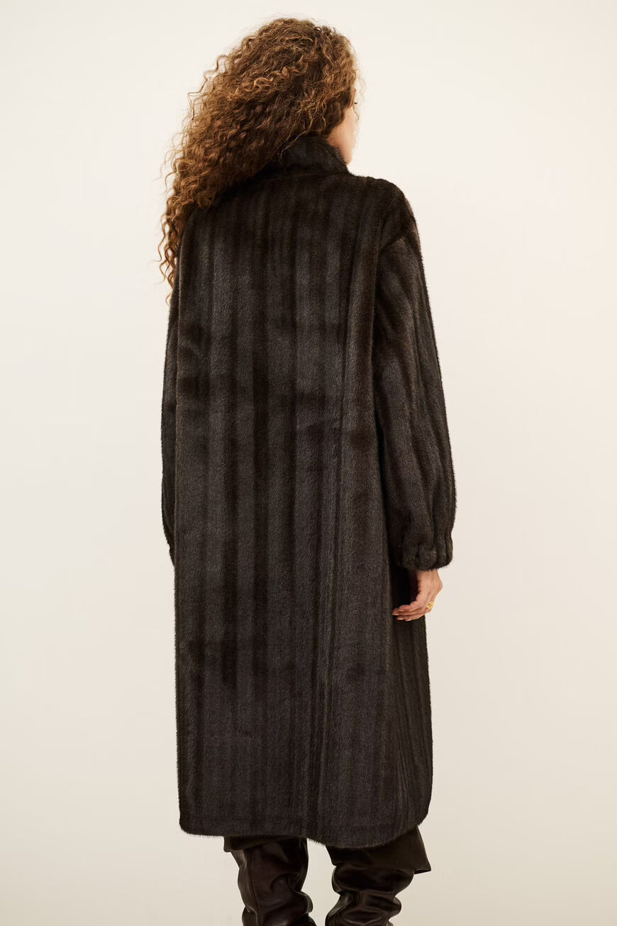 A model viewed from behind wearing a long, dark brown shaggy faux fur coat, black pants, and black boots against a plain off-white background.