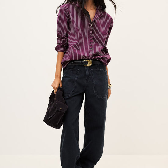 A model stands against a plain light background wearing a purple and black vertically striped button-down shirt, dark wide-leg jeans, black boots, and holding a black handbag.