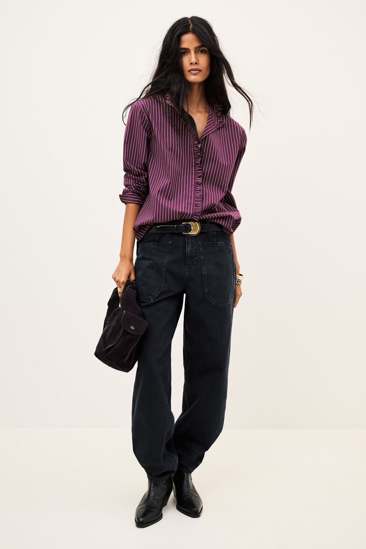 A model stands against a plain light background wearing a purple and black vertically striped button-down shirt, dark wide-leg jeans, black boots, and holding a black handbag.