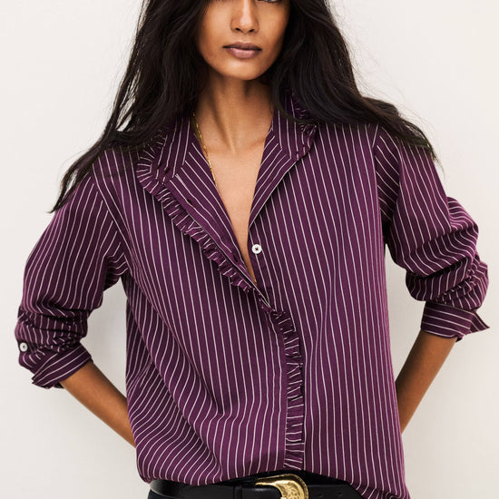 A woman modeling a purple and white vertically striped ruffle blouse with rolled-up sleeves, paired with black jeans and a black belt with a gold buckle, against a plain off-white background.