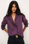 A woman modeling a purple and white vertically striped ruffle blouse with rolled-up sleeves, paired with black jeans and a black belt with a gold buckle, against a plain off-white background.