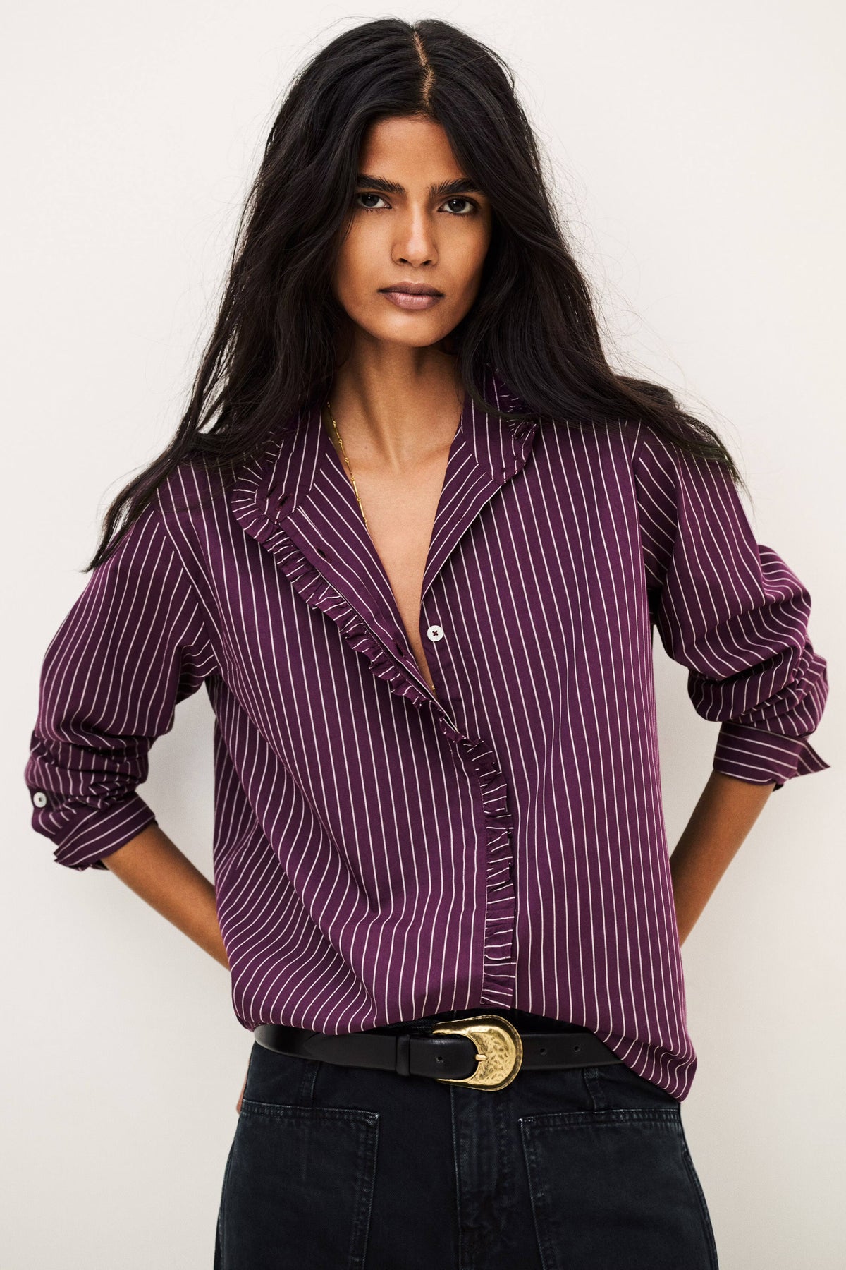 A woman modeling a purple and white vertically striped ruffle blouse with rolled-up sleeves, paired with black jeans and a black belt with a gold buckle, against a plain off-white background.