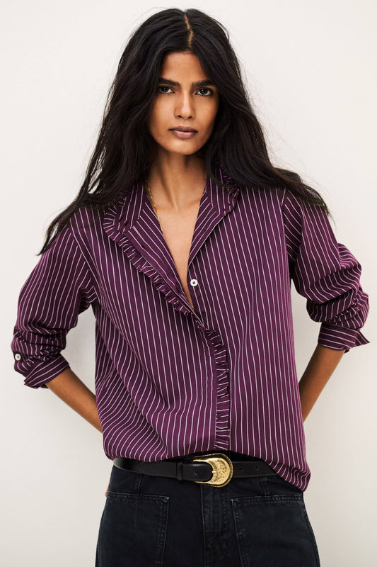 A woman modeling a purple and white vertically striped ruffle blouse with rolled-up sleeves, paired with black jeans and a black belt with a gold buckle, against a plain off-white background.