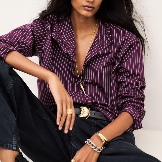 A woman models a purple and white pinstriped button-down shirt with dark wide-leg jeans, a black belt with a gold buckle, and gold jewelry, posed against a light gray background.