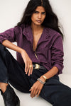 A woman models a purple and white pinstriped button-down shirt with dark wide-leg jeans, a black belt with a gold buckle, and gold jewelry, posed against a light gray background.