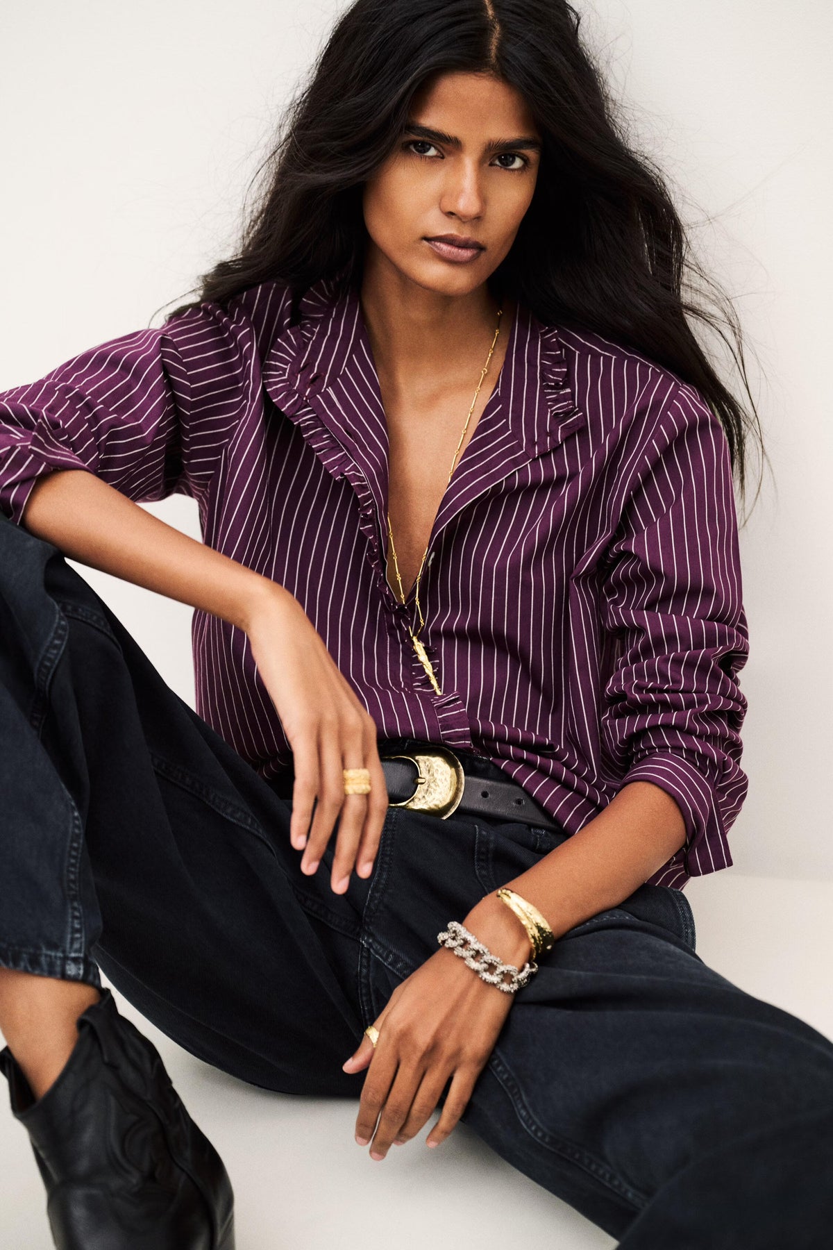 A woman models a purple and white pinstriped button-down shirt with dark wide-leg jeans, a black belt with a gold buckle, and gold jewelry, posed against a light gray background.