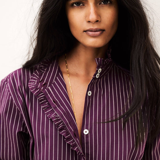 A close-up portrait of a woman wearing a purple and white vertically striped button-up shirt with ruffle details on the collar and front placket, set against a plain light background.