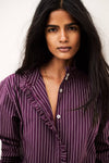A close-up portrait of a woman wearing a purple and white vertically striped button-up shirt with ruffle details on the collar and front placket, set against a plain light background.