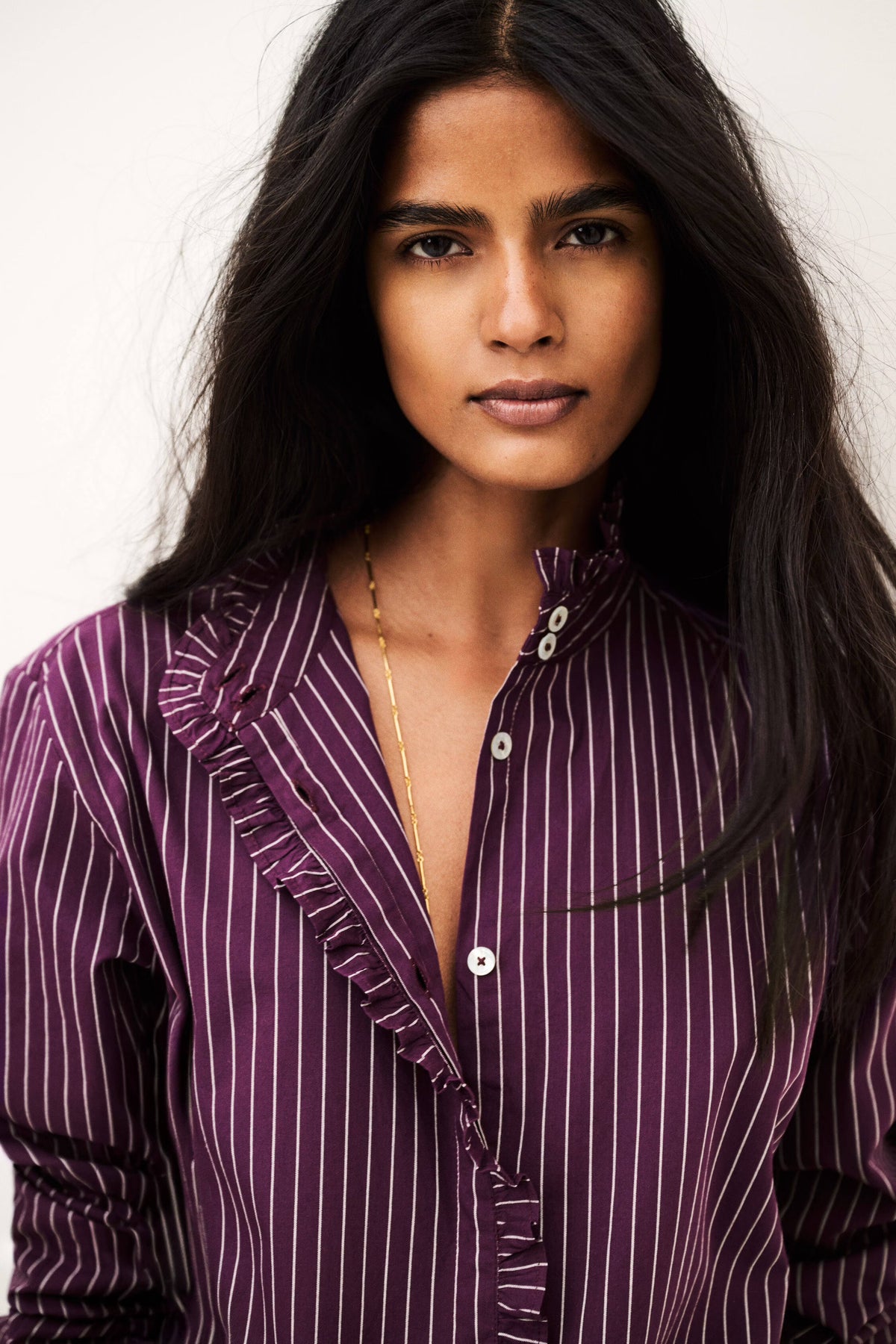 A close-up portrait of a woman wearing a purple and white vertically striped button-up shirt with ruffle details on the collar and front placket, set against a plain light background.