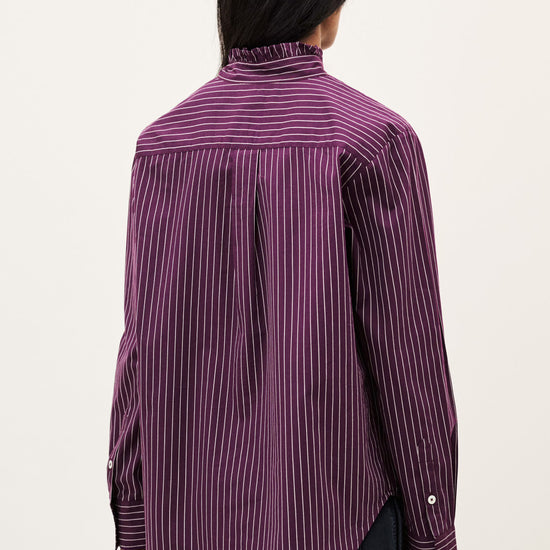 A person shown from the back wearing a purple and black vertically striped button-up shirt with a high mock neck, paired with black pants, against a plain off-white background.