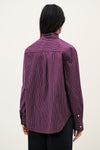 A person shown from the back wearing a purple and black vertically striped button-up shirt with a high mock neck, paired with black pants, against a plain off-white background.