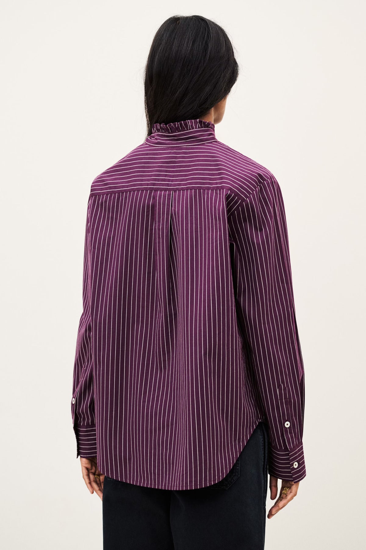 A person shown from the back wearing a purple and black vertically striped button-up shirt with a high mock neck, paired with black pants, against a plain off-white background.