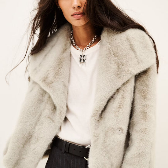 A model wearing a light gray fuzzy faux fur jacket over a white t-shirt and dark pinstripe trousers, photographed against a plain off-white background.