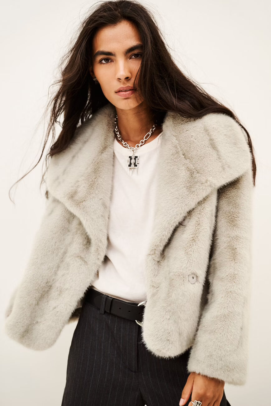 A model wearing a light gray fuzzy faux fur jacket over a white t-shirt and dark pinstripe trousers, photographed against a plain off-white background.