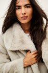 A model wearing a light gray fuzzy faux fur jacket over a white t-shirt and dark pinstripe trousers, photographed against a plain off-white background.