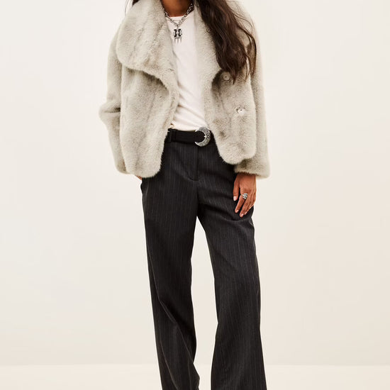 A model wearing a light gray fuzzy faux fur jacket over a white t-shirt and dark pinstripe trousers, photographed against a plain off-white background.