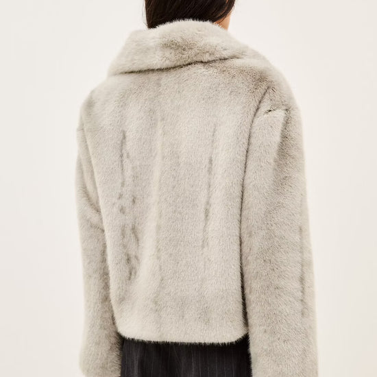 A model wearing a light gray fuzzy faux fur jacket over a white t-shirt and dark pinstripe trousers, photographed against a plain off-white background.