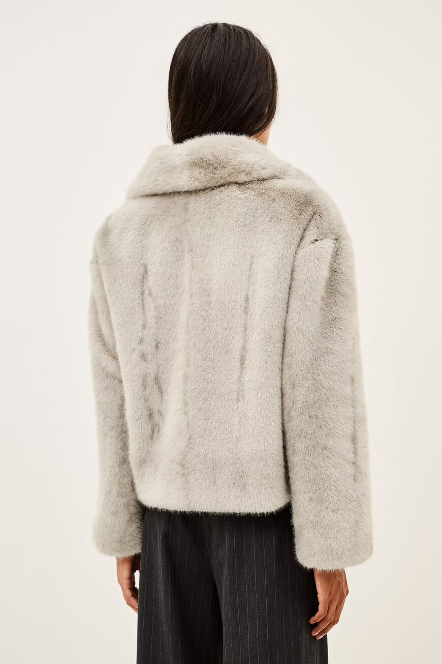 A model wearing a light gray fuzzy faux fur jacket over a white t-shirt and dark pinstripe trousers, photographed against a plain off-white background.