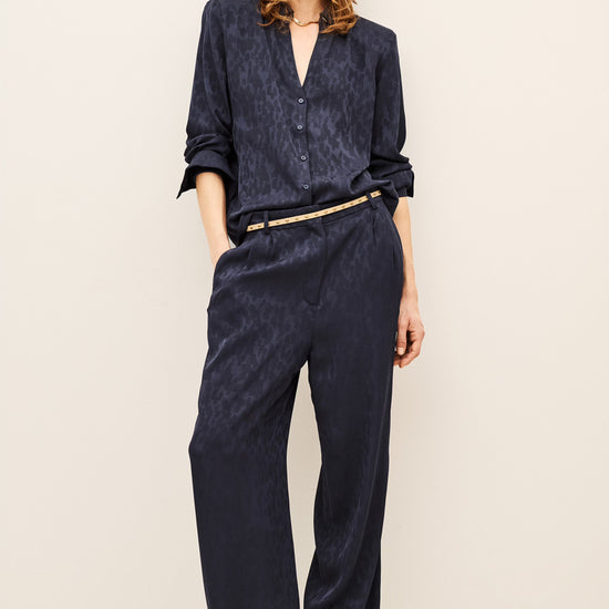 A model wearing a matching navy blue textured two-piece set consisting of a long-sleeve button-up shirt and wide-leg trousers, styled with a thin gold belt and tan suede shoes against a plain off-white background.