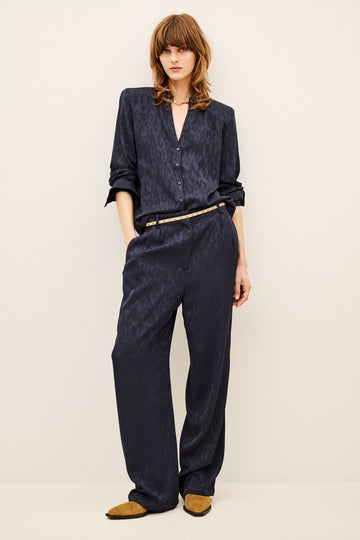 A model wearing a matching navy blue textured two-piece set consisting of a long-sleeve button-up shirt and wide-leg trousers, styled with a thin gold belt and tan suede shoes against a plain off-white background.