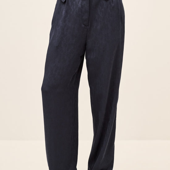 A person wearing dark navy wide-leg trousers with a matching dark navy shirt and tan shoes, shown from the waist down against a plain light beige studio background.