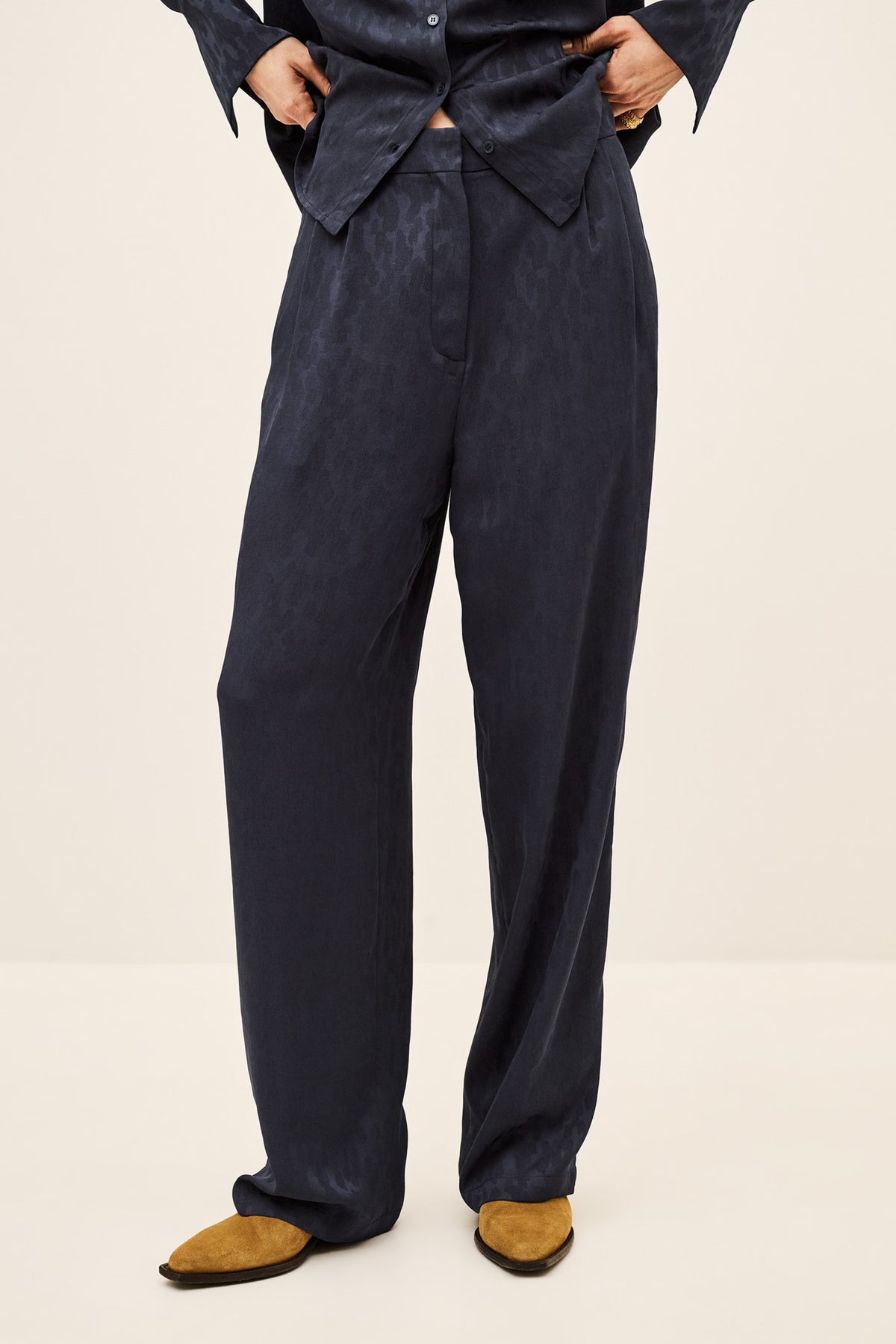 A person wearing dark navy wide-leg trousers with a matching dark navy shirt and tan shoes, shown from the waist down against a plain light beige studio background.