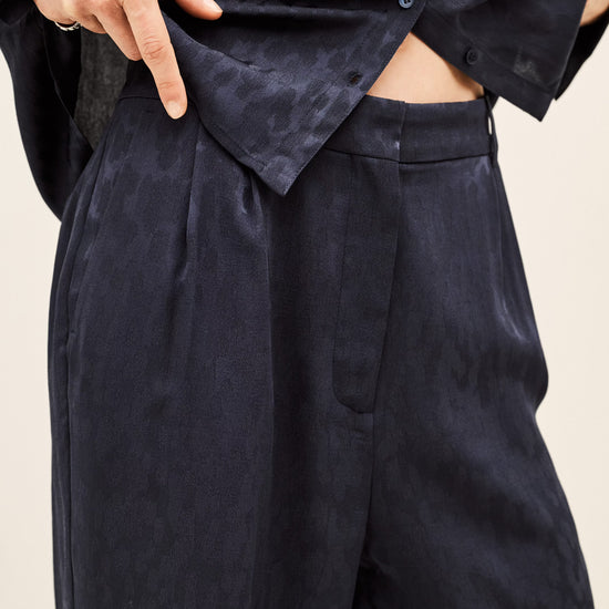 Close-up of a person wearing dark navy wide-leg trousers and a matching dark navy button-up shirt, with hands adjusting the shirt and pulling the fabric to show texture.