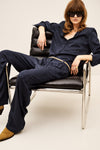A model wearing a dark navy blue textured two-piece set consisting of a long-sleeve button-up shirt and matching wide-leg trousers, styled with a thin gold belt, black sunglasses, and tan suede boots while sitting on a black leather chair with a chrome frame.