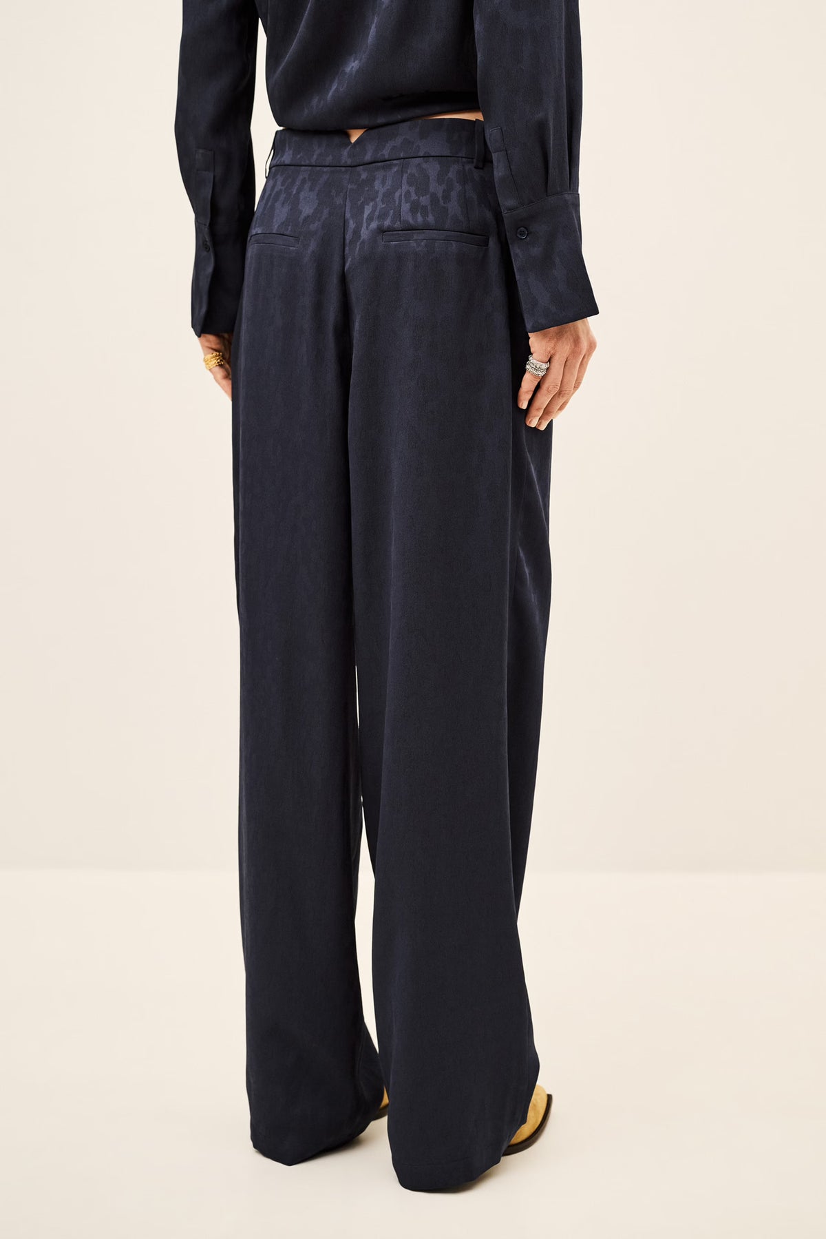 A person shown from the back wearing dark navy wide-leg trousers and a matching dark top against a plain light beige studio background.