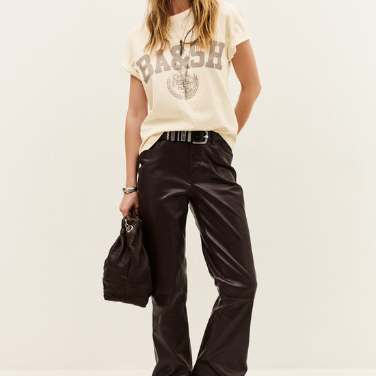 A model stands against a plain off-white studio background wearing a cream graphic t-shirt with 'BASH' and a globe print, paired with shiny black wide-leg pants, a black belt, and holding a black handbag.