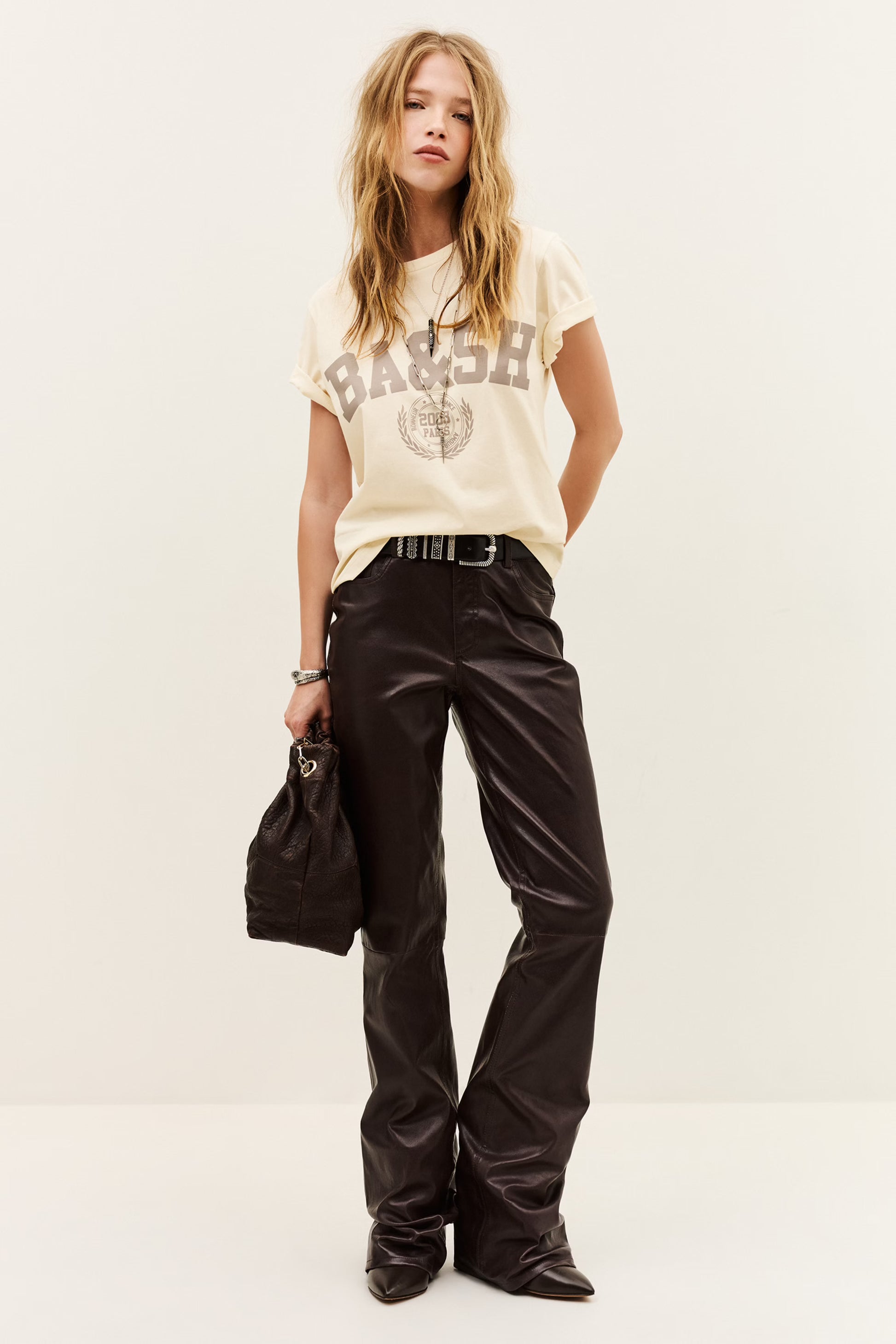 A model stands against a plain off-white studio background wearing a cream graphic t-shirt with 'BASH' and a globe print, paired with shiny black wide-leg pants, a black belt, and holding a black handbag.