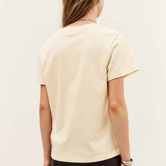 A woman shown from the back wearing a cream-colored short-sleeve t-shirt and black pants, photographed against a plain off-white studio background.