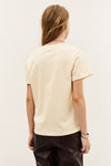 A woman shown from the back wearing a cream-colored short-sleeve t-shirt and black pants, photographed against a plain off-white studio background.